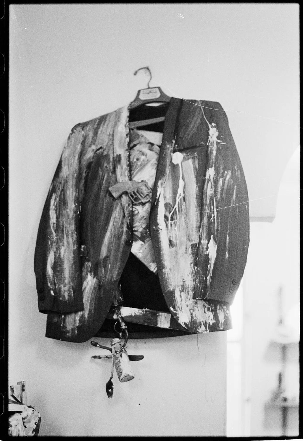 A black-and-white photograph of a paint-splattered jacket hanging on a hook against the wall