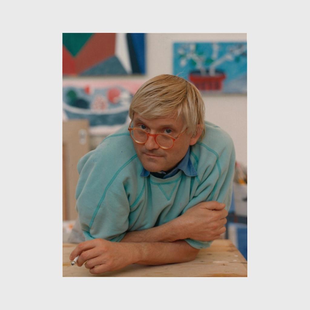 Hockney in a turquoise jumper and round glasses in his studio