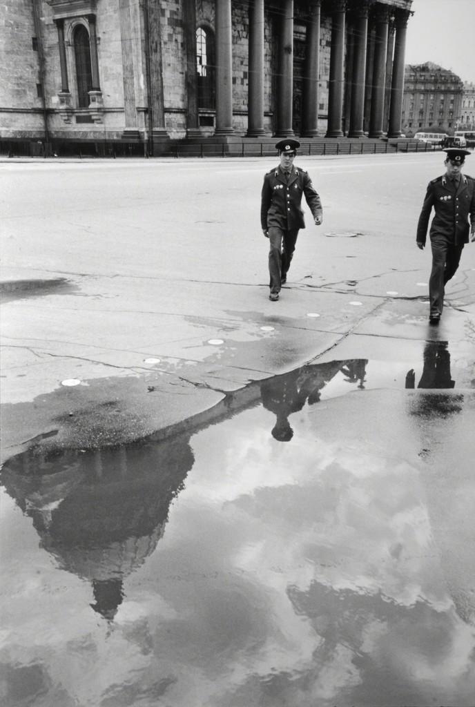 Two uniformed soldiers walk along a colonnade, their silhouettes reflected in a puddle on the square