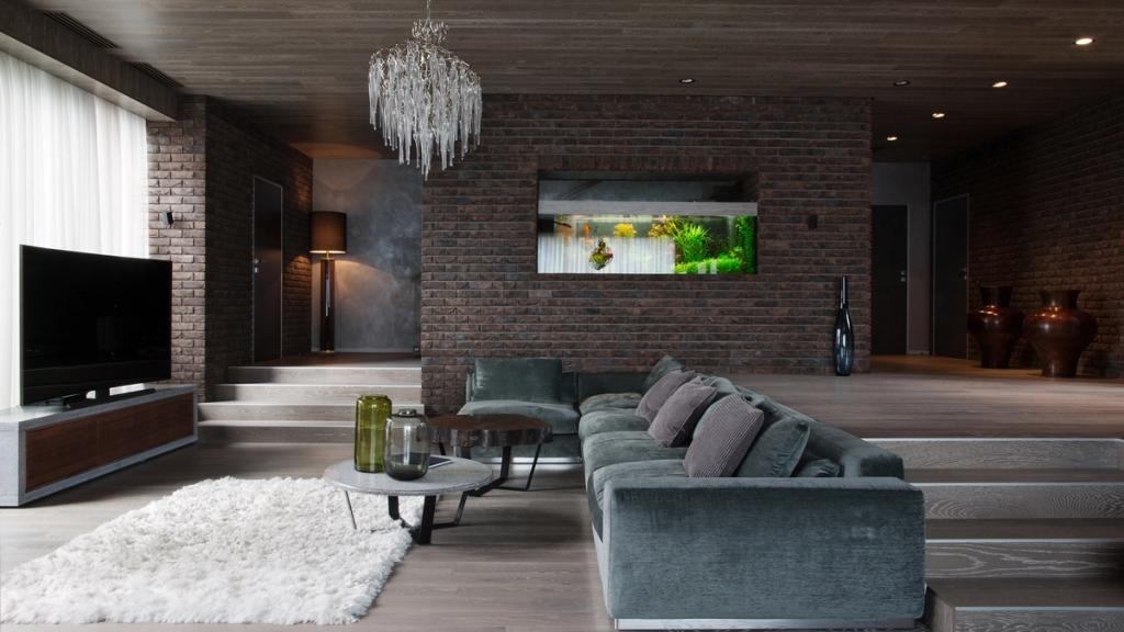 A spacious living room with brick walls, a grey sofa, a crystal chandelier and a built-in aquarium