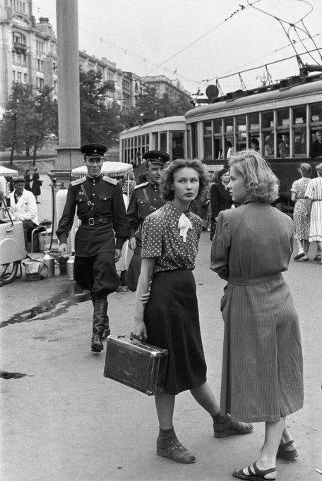 Two young women talk on the street; Soviet officers pass behind them near a tram