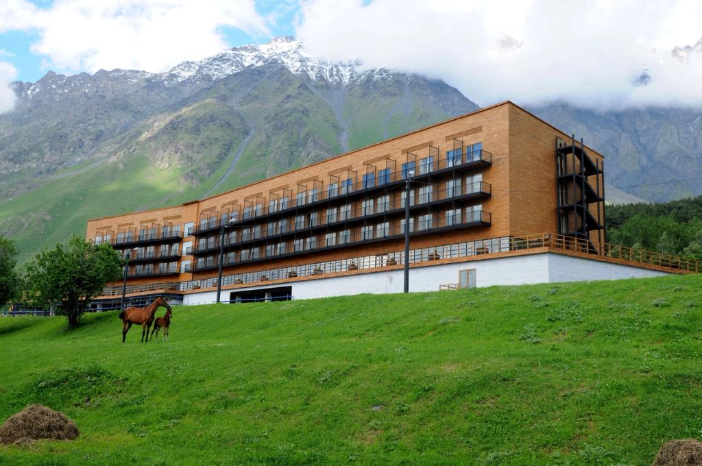 The timber wing of Rooms Hotel Kazbegi against a backdrop of snow-capped Kazbegi mountains, with horses grazing in the meadow