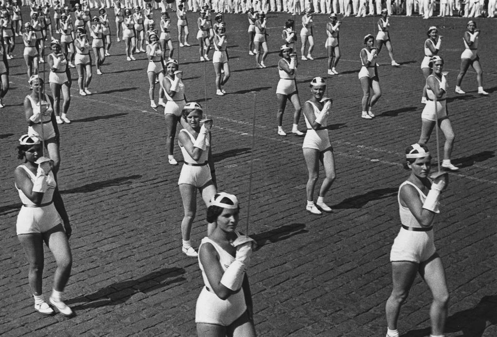 A physical-culture parade: rows of young women in white uniforms perform exercises on cobblestones