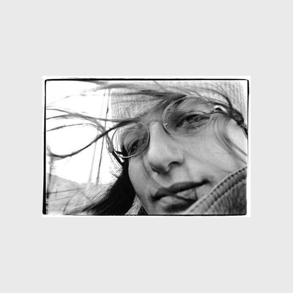 A black-and-white close-up portrait of a young woman in glasses, her hair blown loose by the wind
