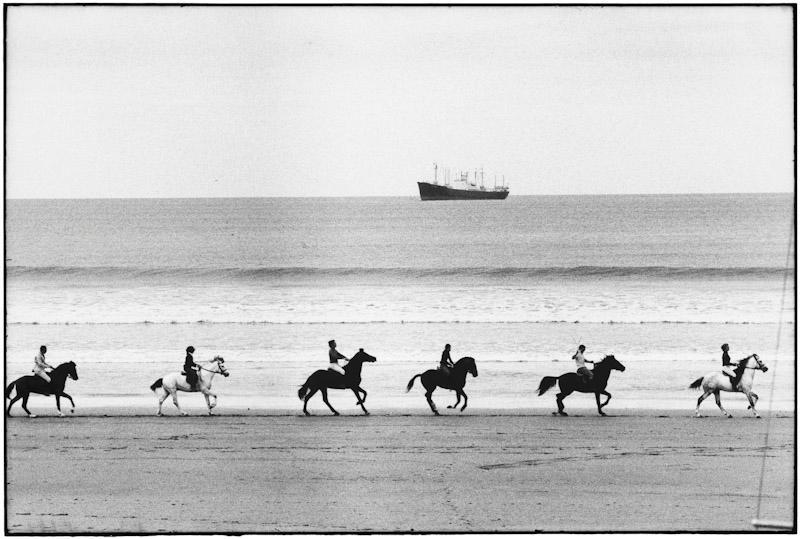 Six riders travel in single file along the water's edge of a beach, a cargo ship on the horizon