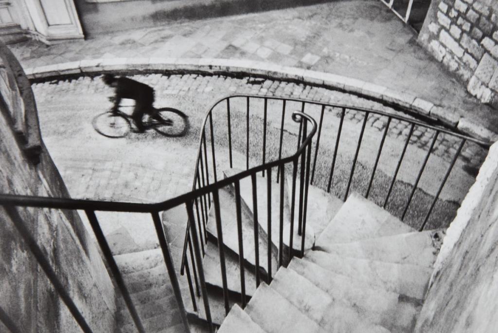A black-and-white photograph of a cyclist on a cobbled street, shot from above a spiral staircase with a wrought-iron railing