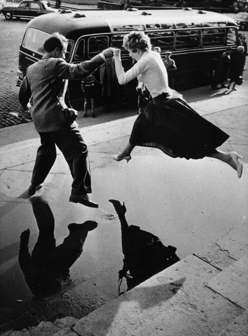 A man and a woman leap over a puddle beside a bus, their reflections visible in the water