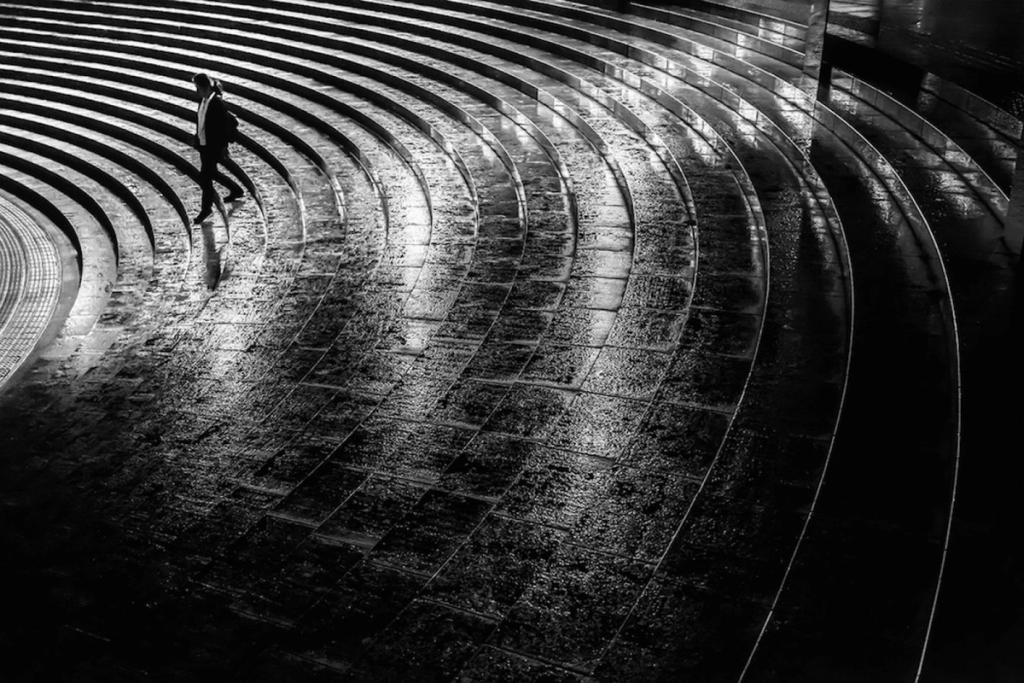 A passerby walks along wet, curved amphitheatre steps in sharp contrasting light