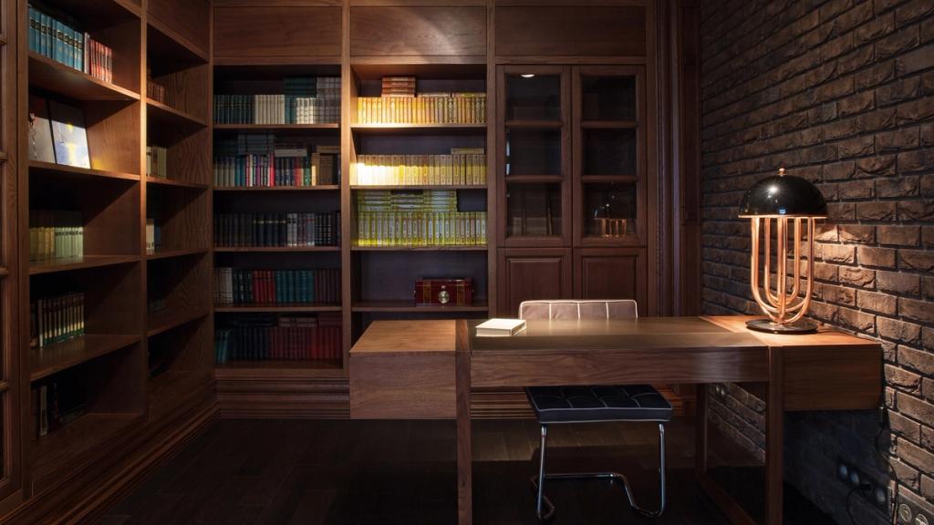 A study with wooden bookshelves, a writing desk and a desk lamp set against a brick wall
