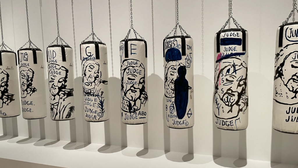 Painted boxing bags bearing faces and the inscription JUDGE, hung in a row in a gallery
