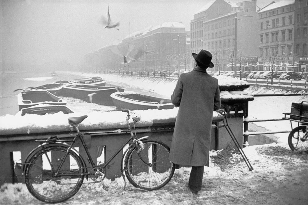 A man in a coat and hat stands beside a bicycle, looking out over a snow-covered embankment lined with barges