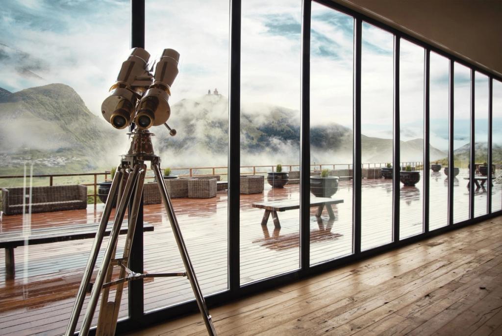 A pair of binoculars on a tripod by a panoramic window, with a terrace and mountain mist beyond