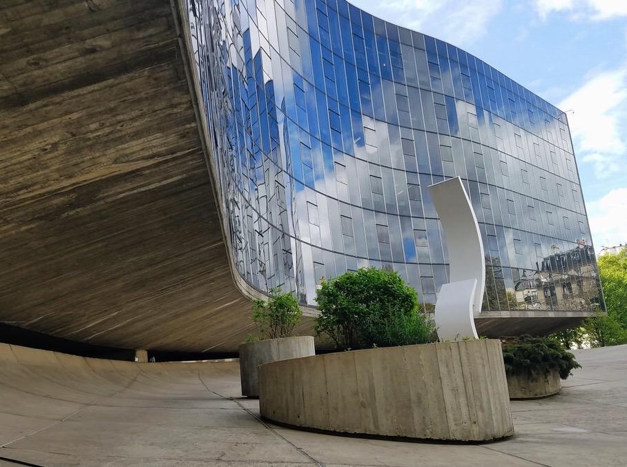 A glass building with a curved concrete canopy and circular flowerbeds in the courtyard