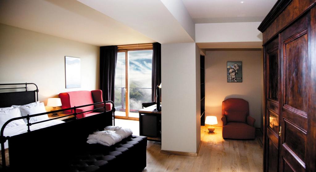A room with a bed, a red sofa and a panoramic window overlooking cloud-wreathed mountains