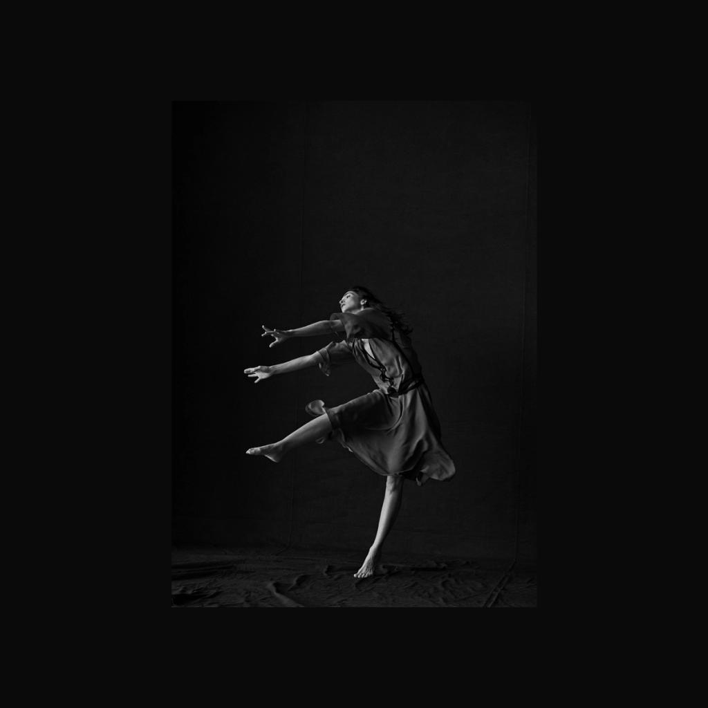 A black-and-white photograph of a woman dancing in mid-leap against a dark background