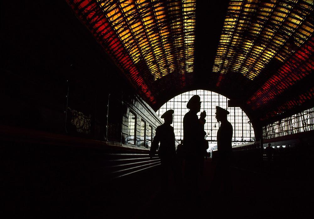 Silhouettes of military figures beneath a curved stained-glass vault of a railway station in red and yellow tones