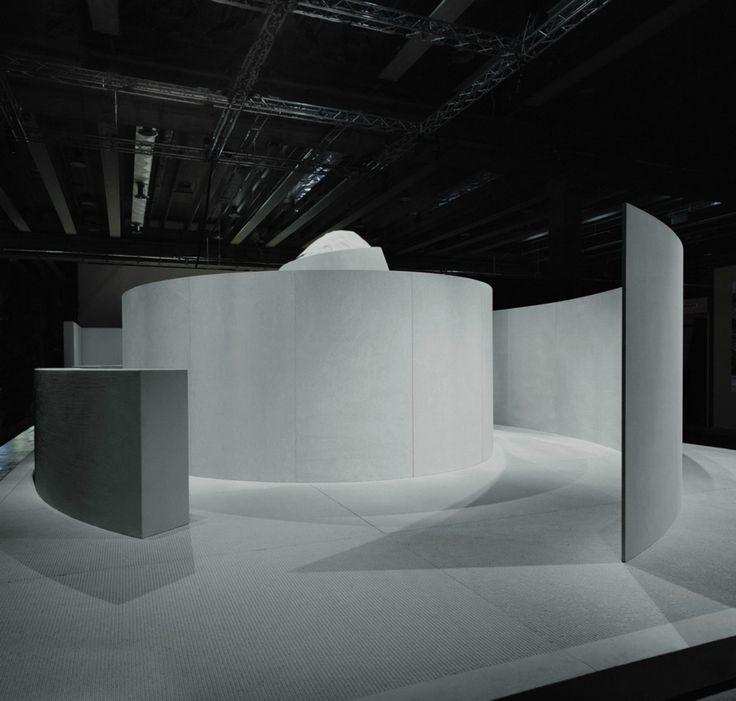 A white cylindrical installation with curved walls beneath the industrial ceiling of an exhibition hall