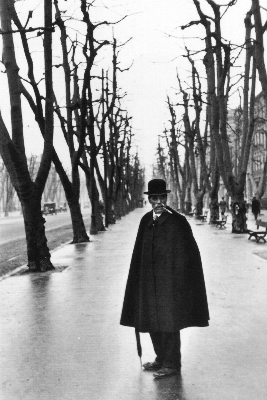 A man in a bowler hat and overcoat, carrying a cane, stands in the middle of an avenue of bare trees