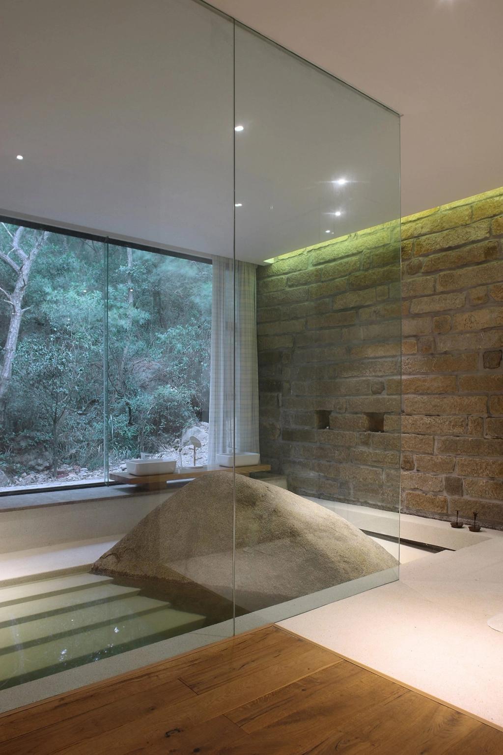 A plunge pool with a natural boulder beside a panoramic window and a backlit stone wall