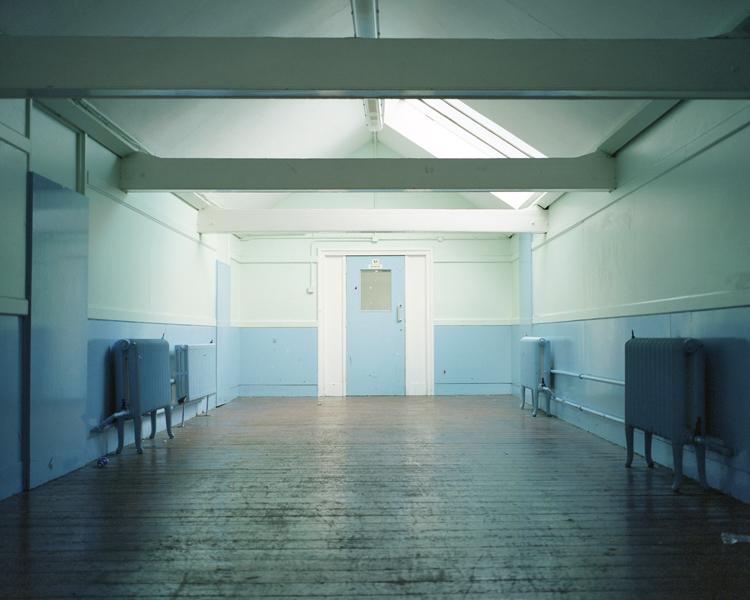 An empty, light-filled corridor with wooden floors and old cast-iron radiators under a faint bluish light