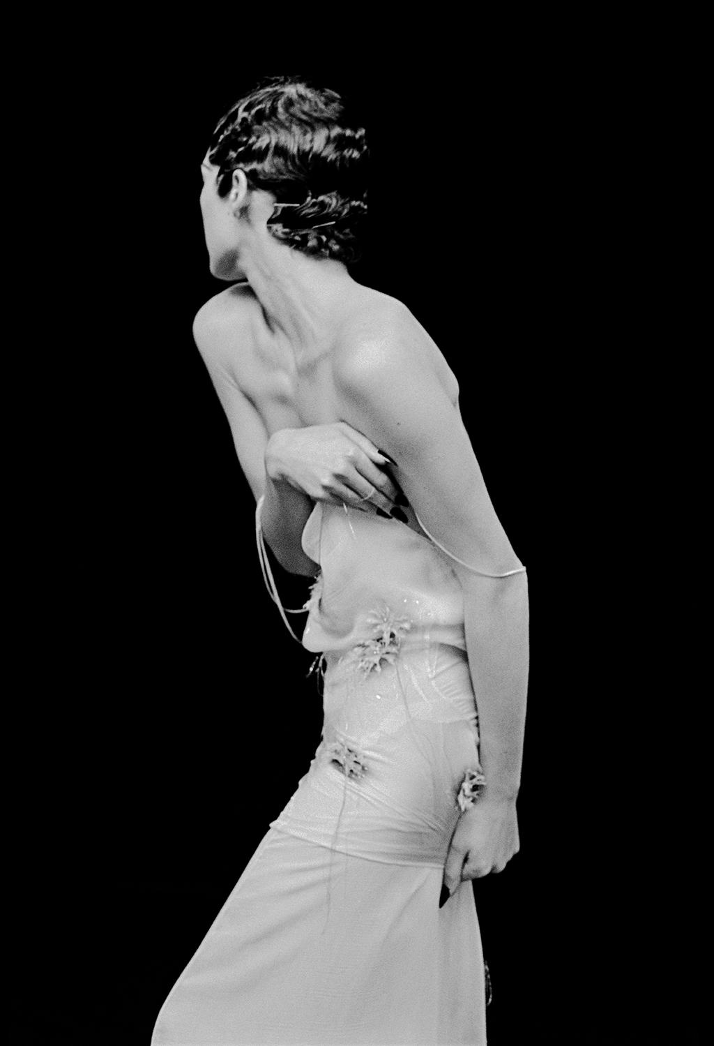 A black-and-white portrait of a model seen from behind, wearing a light dress against a dark background