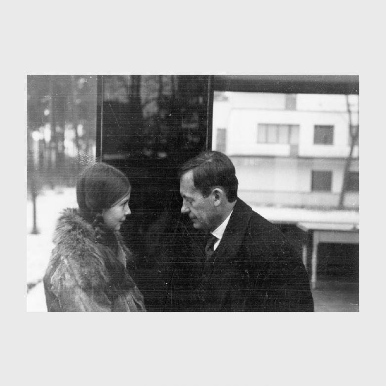 Black-and-white photograph: a man in an overcoat talks to a young girl in a fur coat on a winter street