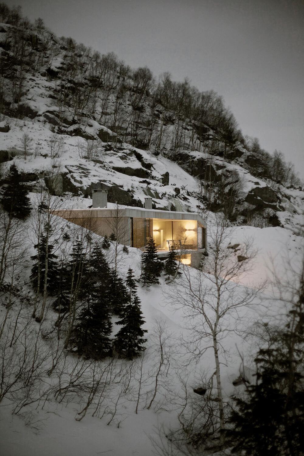 A house cut into a snowy mountain slope, its windows glowing warmly in the dusk