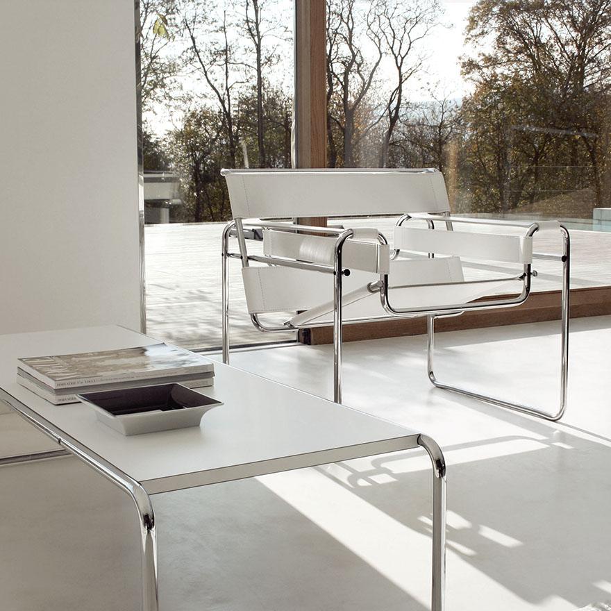 A white Wassily Chair by a panoramic window, beside a low table and books