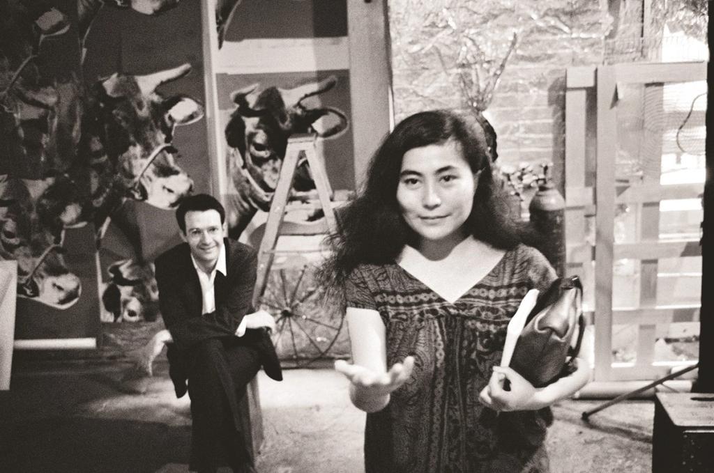 Black-and-white photograph in the studio: a woman extends her arm while a man sits in front of works depicting cows