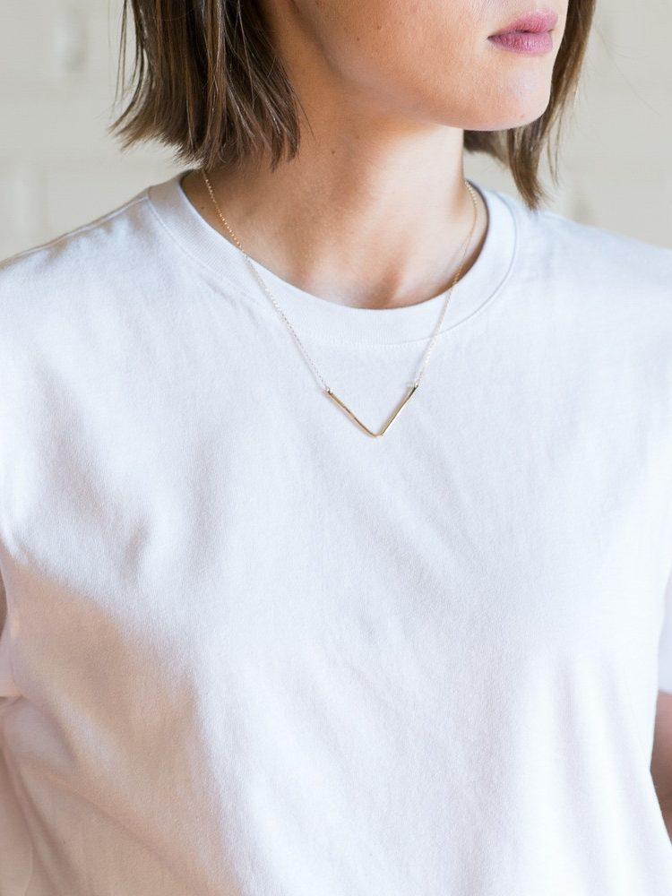 A gold V-pendant necklace on the neck of a woman in a white T-shirt