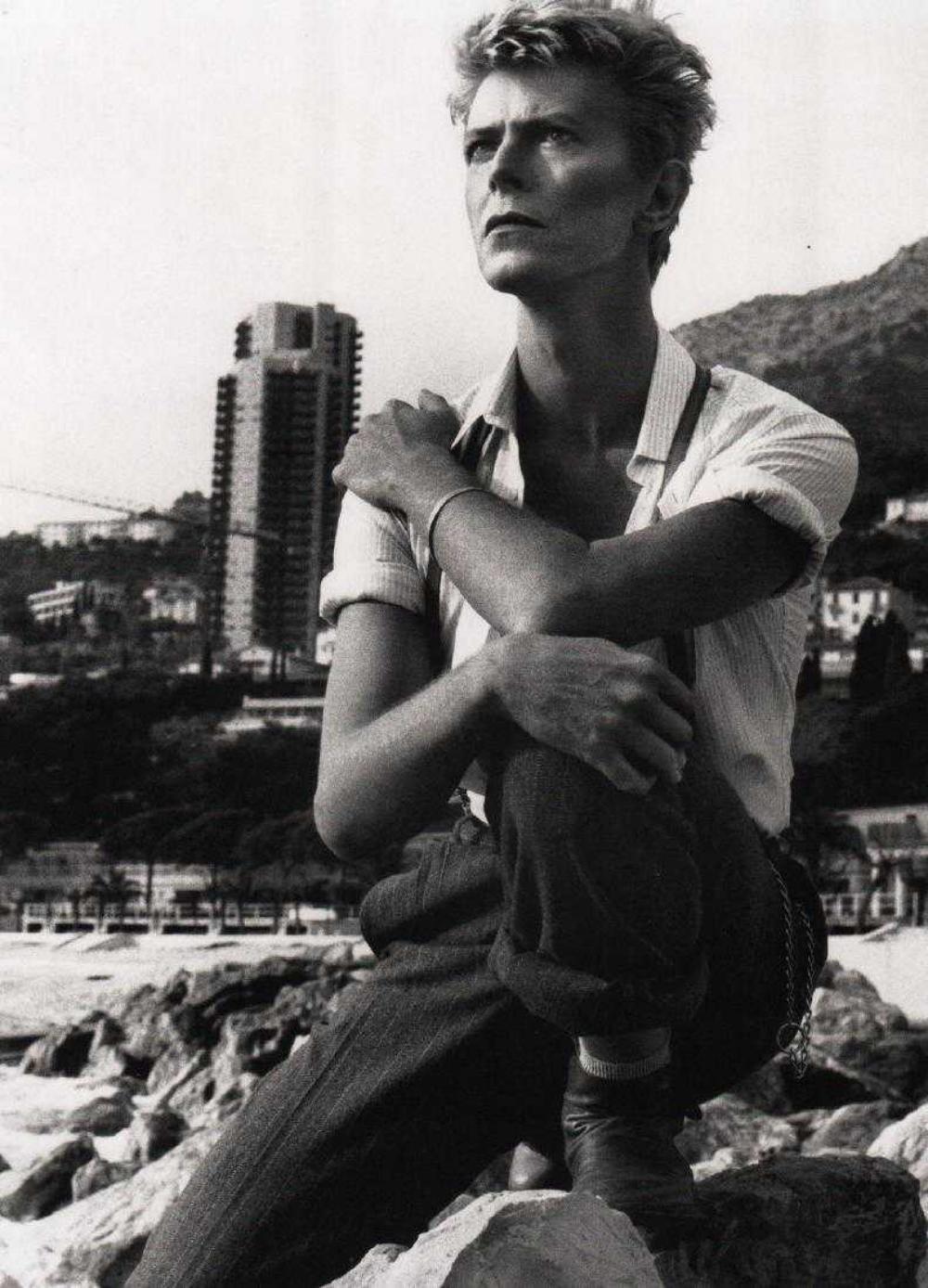 A fair-haired man sits on coastal rocks with a high-rise building and mountains behind him, black-and-white portrait