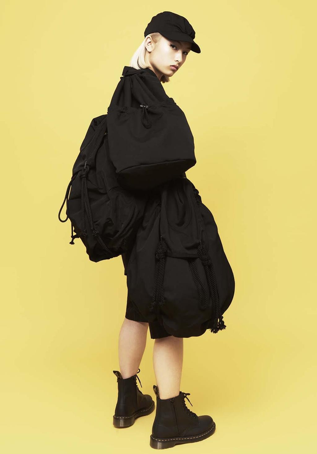 A model in profile carrying two black drawstring backpacks, with a cap and boots