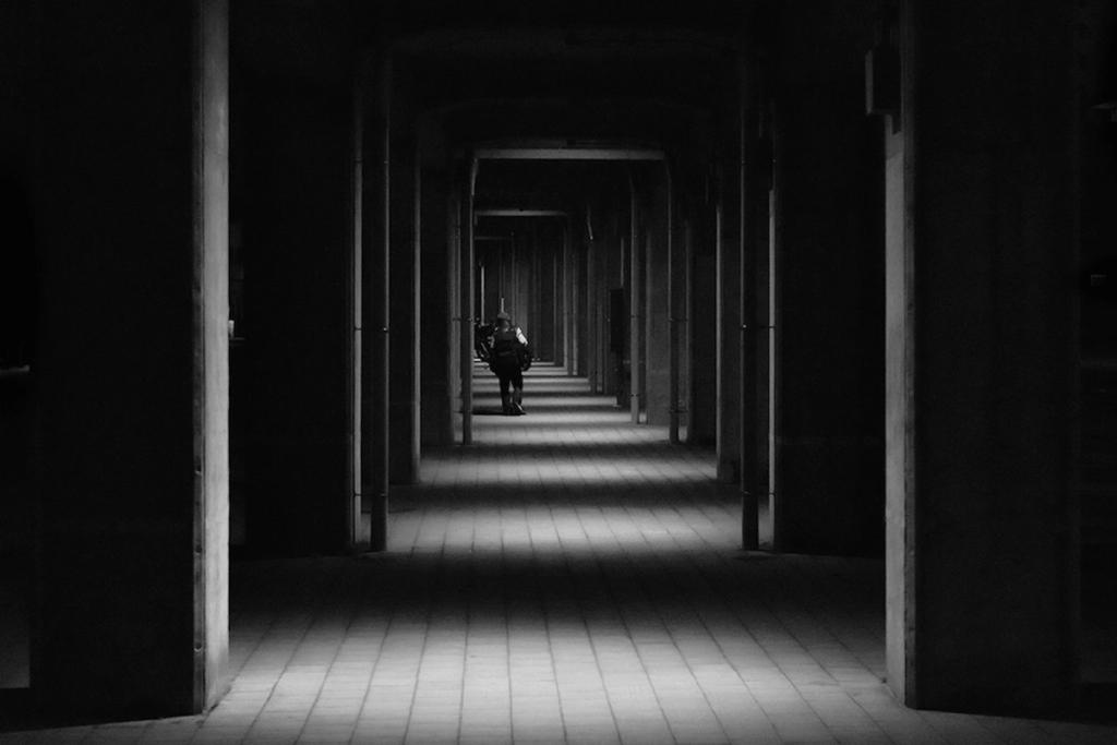 A dark covered arcade with rows of columns; a solitary figure walks in the distance
