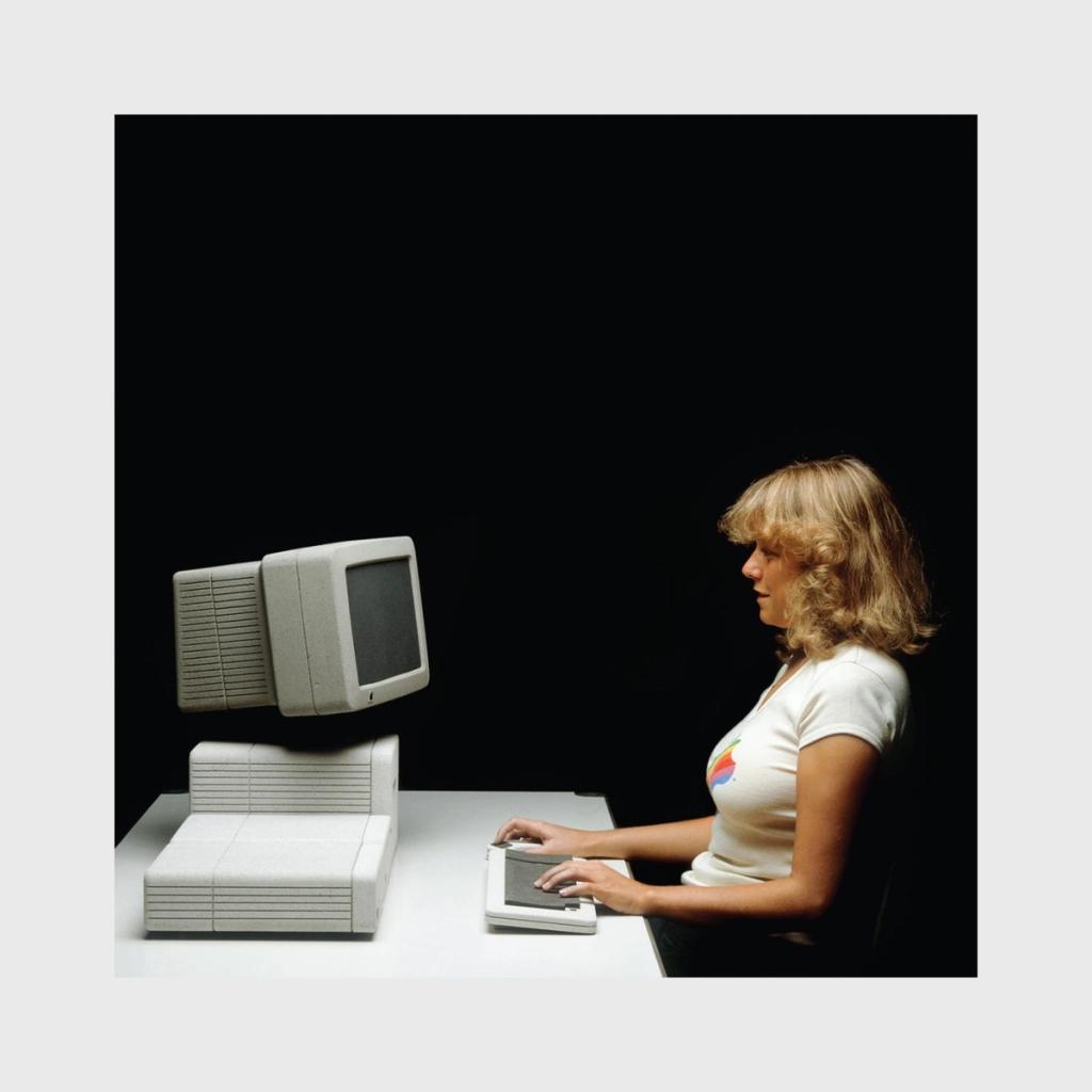A woman at a white Apple desktop computer wearing a T-shirt with the multicoloured Apple logo