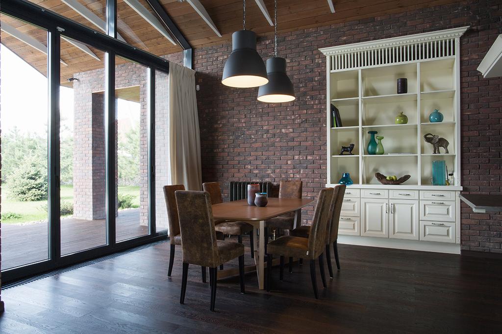 Dining area with a wooden table and chairs against a brick wall, a white open shelving unit and panoramic windows overlooking the garden