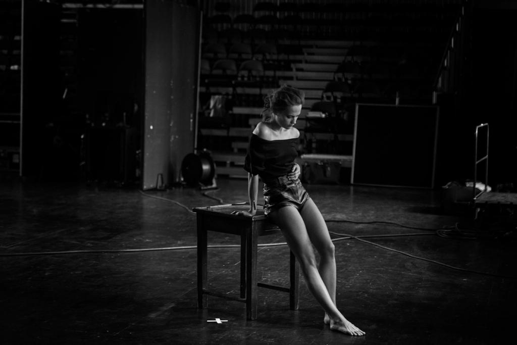 A woman sits on a wooden table on an empty stage in half-darkness