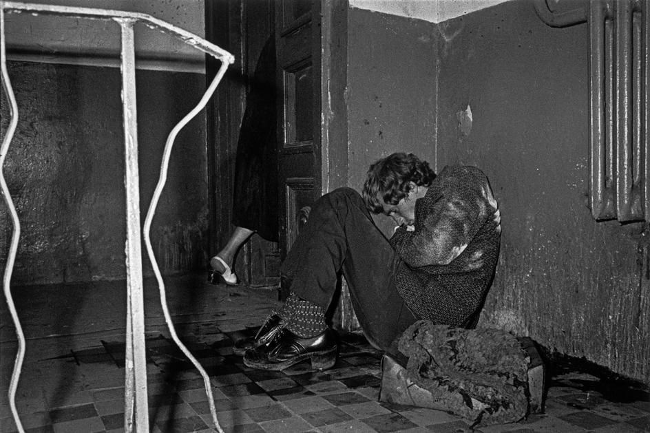 A young man sits on the floor in the corner of a dilapidated room, hugging his knees