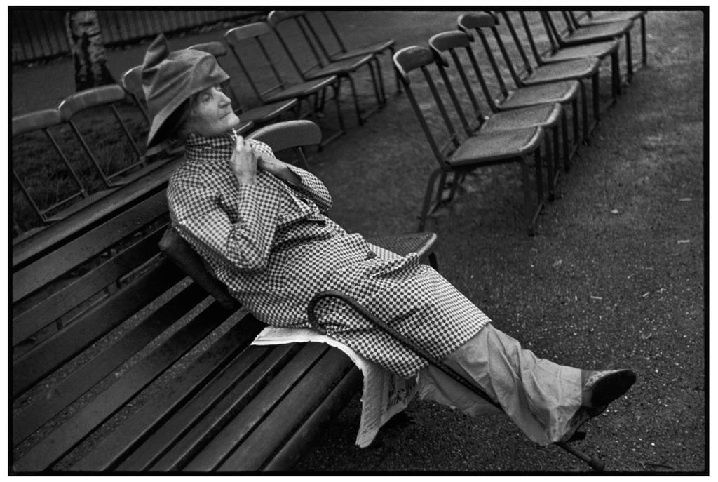 An elderly woman in a hat reclines on a bench amid empty chairs in a park