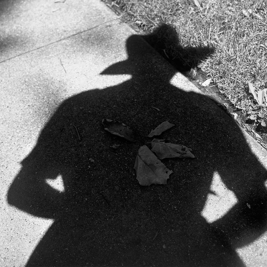 Black-and-white self-portrait by photographer Vivian Maier as a shadow wearing a hat, cast on a pavement