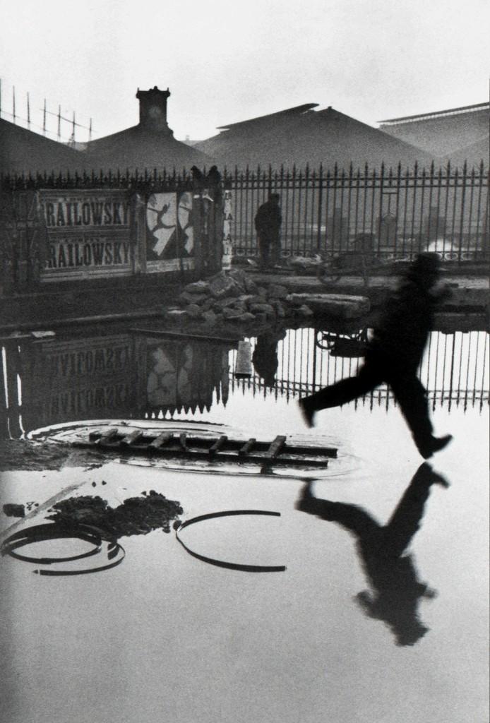 A black-and-white frame by Henri Cartier-Bresson: a man leaping over a flooded square near the Gare Saint-Lazare