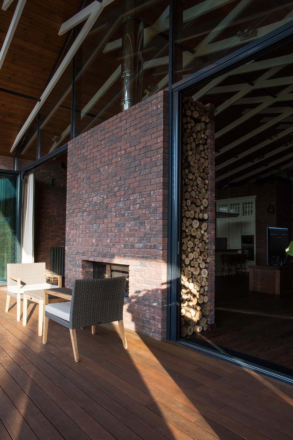 A brick see-through fireplace with a side log store on a terrace with wooden decking and floor-to-ceiling glazing