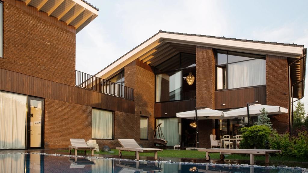A brick house with timber inserts, a balcony and parasols beside a pool with sun loungers