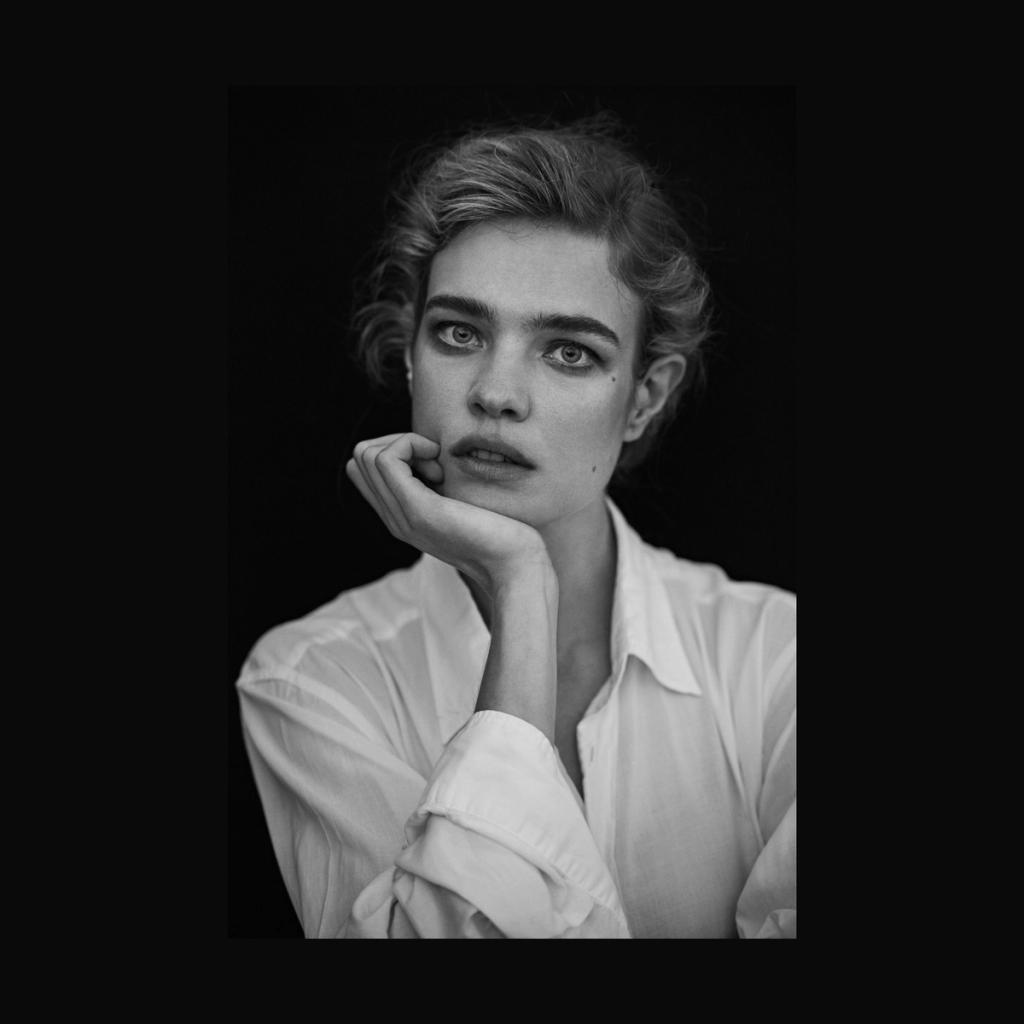 A black-and-white portrait of a young woman in a white shirt, resting her chin on her hand