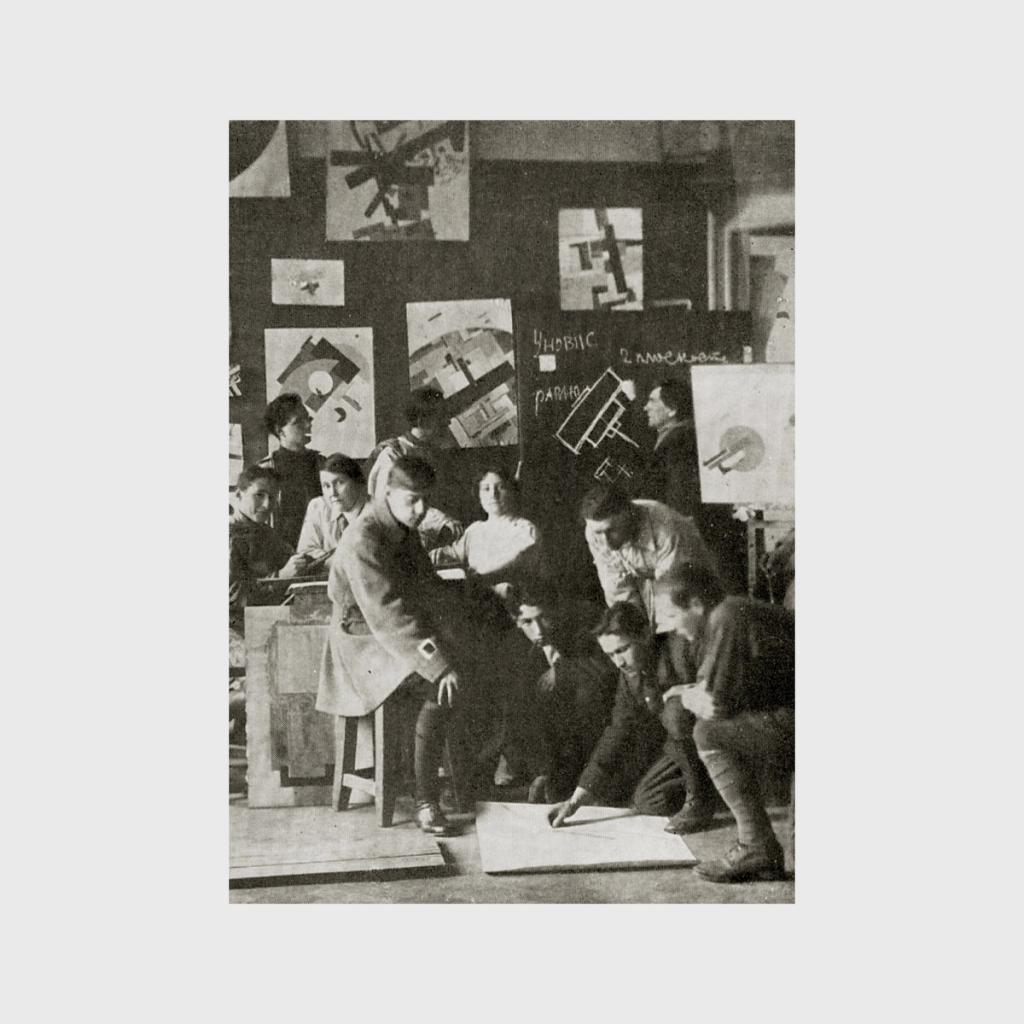 A black-and-white photograph of Malevich's students at work against a backdrop of Suprematist sheets and the UNOVIS inscription