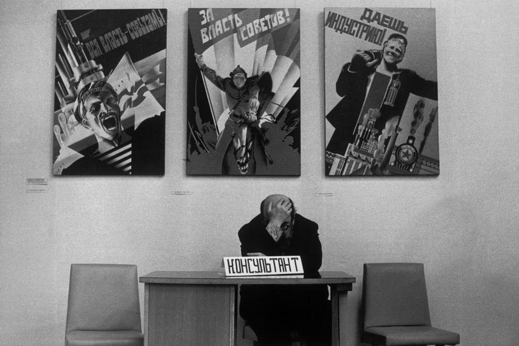 A man covers his face with his hand at a table beneath Soviet propaganda posters