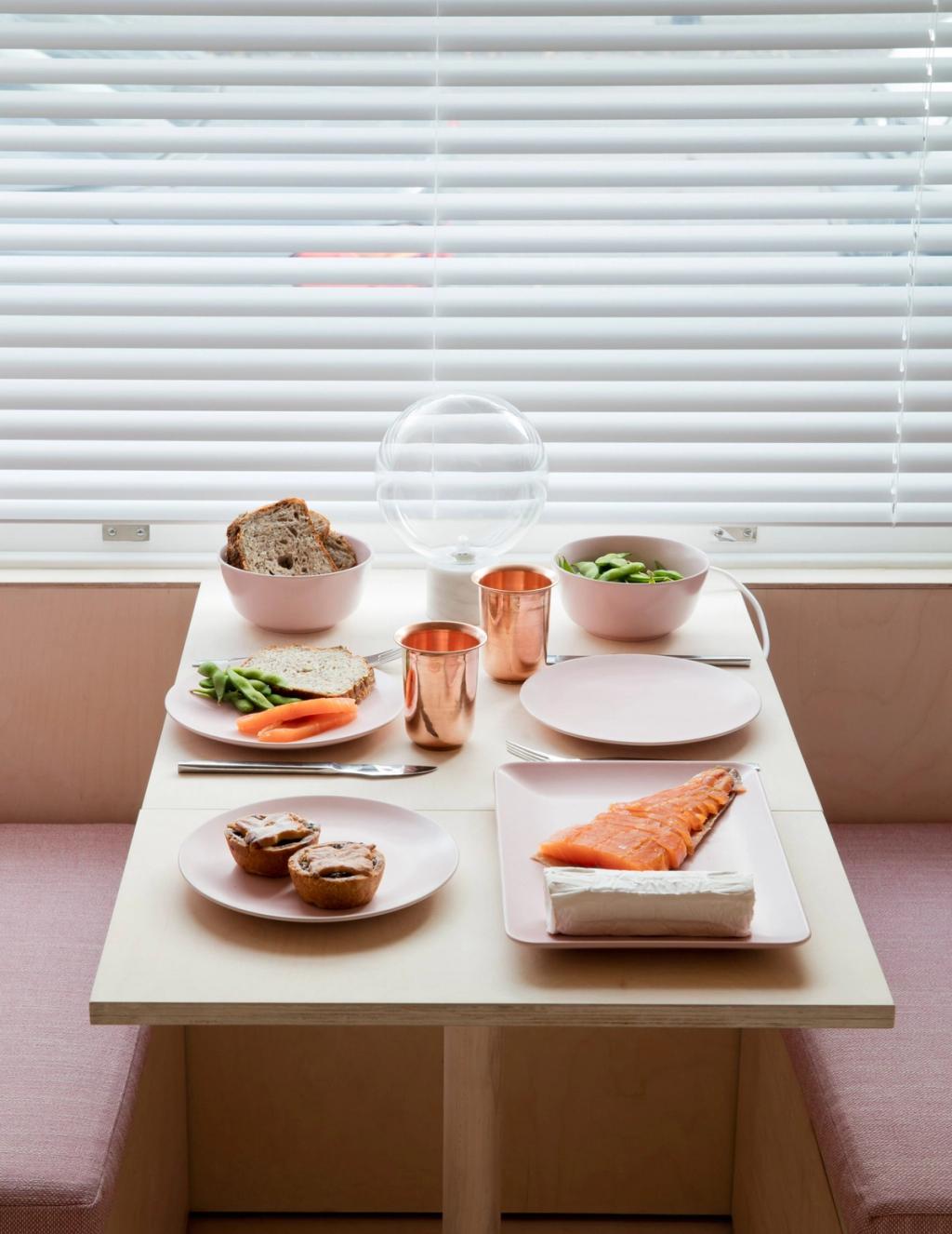 Накрытый обеденный стол с розовой посудой, медными стаканами, хлебом и лососем у окна с жалюзи