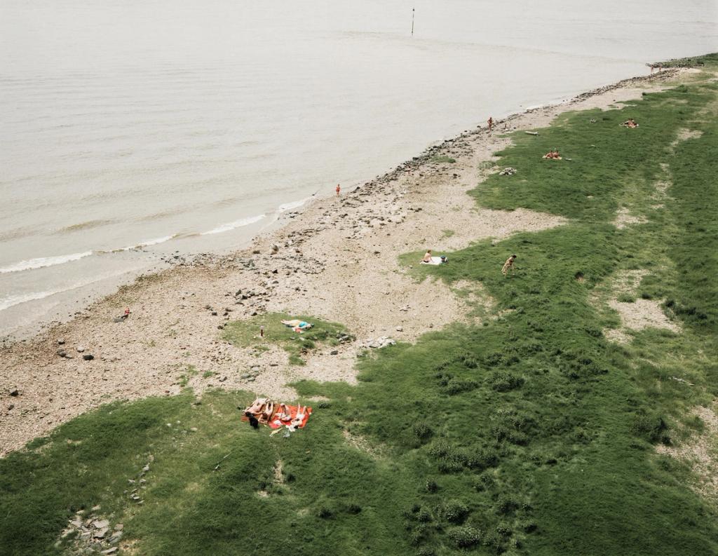 A sandy bank of a wide river with scattered holidaymakers on a strip of green grass