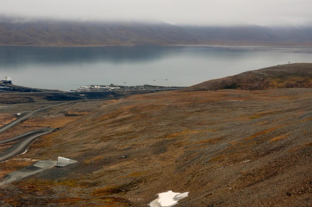 A panoramic view of a Svalbard bay with harbour structures and a gently sloping expanse of brown tundra beneath the clouds
