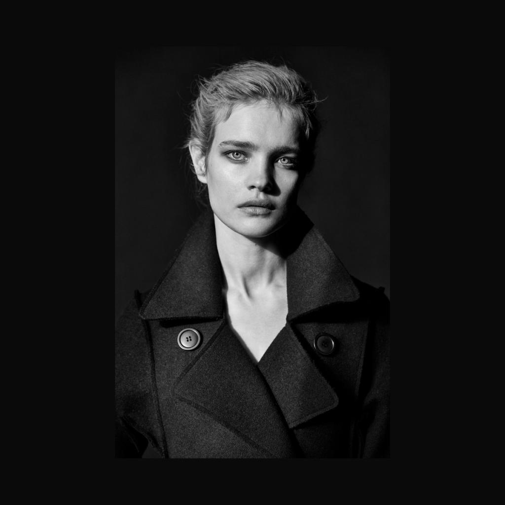 A black-and-white portrait of a fair-haired woman in a dark coat against a black background