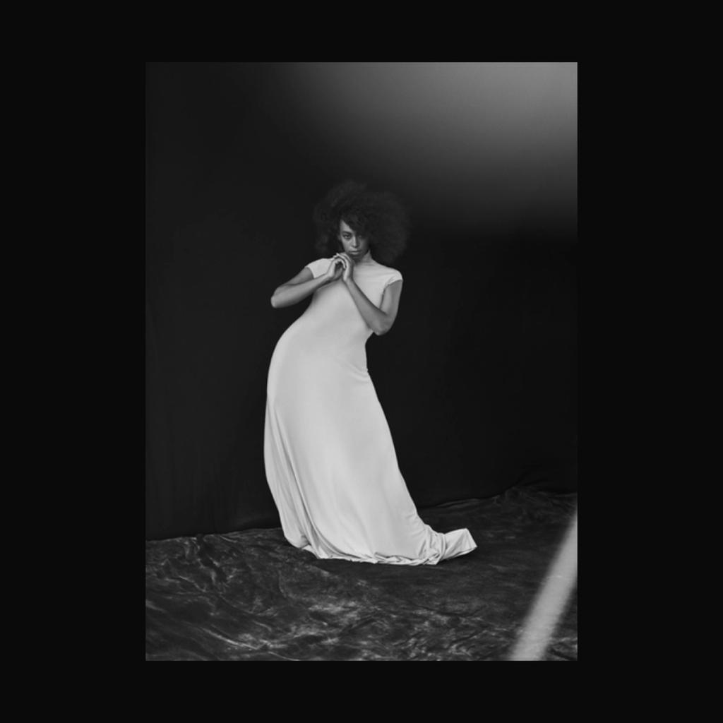 A woman in a long light-coloured dress stands against a dark draped background